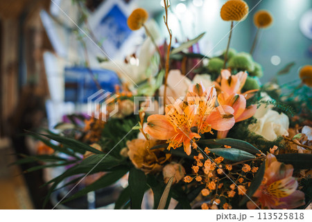 Colorful floral arrangement featuring vibrant orange lilies, small orange flowers, yellow button like blooms, and varied greenery in a blurry indoor setting. Colorful floral arrangement featuring vibrant orange lilies, small orange flowers, yellow button like blooms, and varied greenery in a blurry indoor setting. 113525818