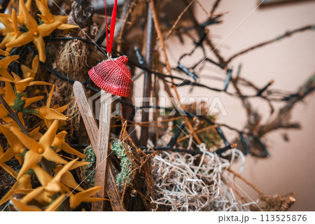 Close up view of textured red knitted hat hanging from ribbon, surrounded by dried plants, twigs, and yellow star shaped flowers in rustic, natural themed arrangement. Indoor display. 113525876