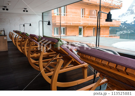 Wooden lounge chairs with purple cushions, black floor lamps, and snowy mountain views in a cozy modern interior at Hotel Eiger in a cold alpine region. Wooden lounge chairs with purple cushions, black floor lamps, and snowy mountain views in a cozy modern interior at Hotel Eiger in a cold alpine region. 113525881