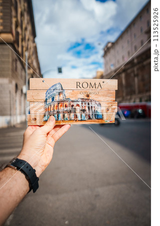 Hand holding souvenir sign with Colosseum image in Rome. ROMA and Colosseo displayed. Blurred street scene background. Urban setting, partly cloudy. 113525926