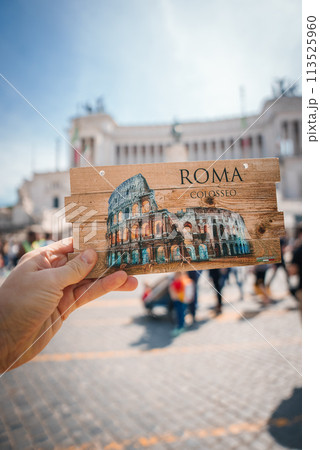 Hand holding Colosseum illustration postcard with ROMA and COLOSSEO in Rome, Italy. Blurred urban background aligns with street architecture, sunny day vibes. 113525960