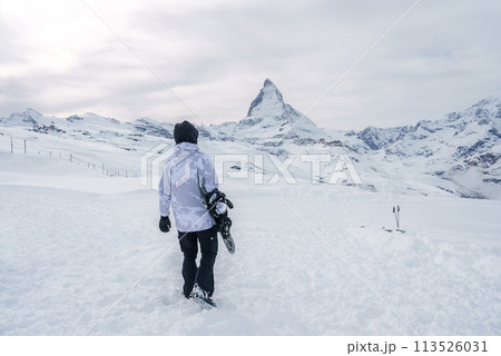 Snowboarder in camouflage winter gear walking towards Matterhorn peak in Zermatt ski resort, Switzerland. Serene snow covered landscape with iconic mountain silhouette. 113526031