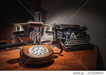 Vintage rotary dial telephone and classic typewriter on polished wooden surface evoke elegance and nostalgia. Luxurious decor in Zermatt, Swiss ski resort. Vintage rotary dial telephone and classic typewriter on polished wooden surface evoke elegance and nostalgia. Luxurious decor in Zermatt, Swiss ski resort. 113526054