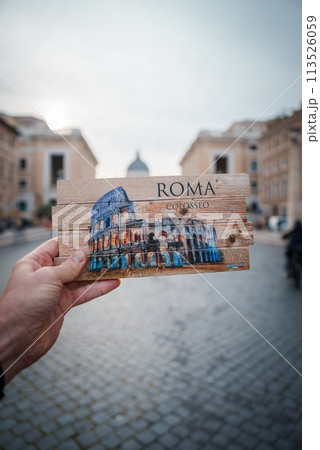 Hand holding postcard with Colosseum illustration, ROMA at top, COLOSSEO below. Urban background resembles Rome, Italy. Sunset or sunrise lighting. Juxtaposition of souvenir and city. 113526059