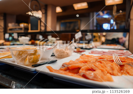 Buffet spread with citrus dessert and sliced salmon on white plate in warm, inviting restaurant or dining area with out of focus background dishes and bar area. 113526169