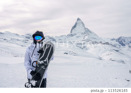 Snowboarder in winter gear holding black snowboard in snowy landscape near Zermatt, Switzerland. Matterhorn mountain in background under overcast sky. Snowboarder in winter gear holding black snowboard in snowy landscape near Zermatt, Switzerland. Matterhorn mountain in background under overcast sky. 113526185