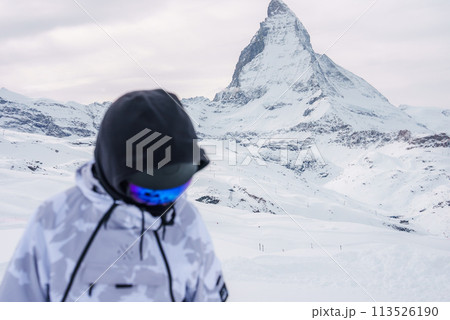 Person in winter sports gear gazes at snowy Matterhorn mountain, a landmark of Zermatt ski resort in Switzerland. Captures alpine winter sports adventure. 113526190