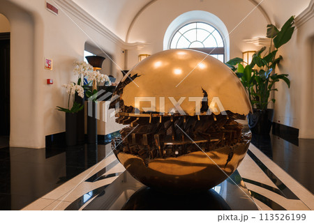 Luxury hotel lobby in Rome featuring large cracked gold sphere sculpture on black pedestal, geometric floor tiles, arched windows, and lush green plants. Blend of art and architecture. 113526199