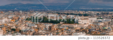 An aerial view of Rome shows a dense cityscape, terracotta roofs, and mixed architecture under a cloudy sky. Its rich history is seen in the mix of old and new buildings, nestled among hills. An aerial view of Rome shows a dense cityscape, terracotta roofs, and mixed architecture under a cloudy sky. Its rich history is seen in the mix of old and new buildings, nestled among hills. 113526372