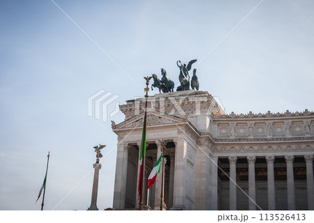 Neoclassical building in Rome with grand columns, bronze sculptures, and Italian flags fluttering under a clear sky. Iconic monument honoring Victor Emmanuel II. Neoclassical building in Rome with grand columns, bronze sculptures, and Italian flags fluttering under a clear sky. Iconic monument honoring Victor Emmanuel II. 113526413