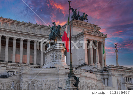 Victor Emmanuel II National Monument in Rome, Italy, bathed in soft sunlight. Italian flag in foreground, statues, columns, tourists, clear sky, golden hour. 113526497