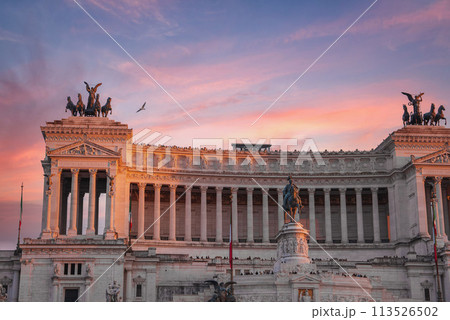 A majestic view of Altare della Patria, or National Monument to Victor Emmanuel II, in Rome, Italy. White marble facade, statues, and Italian flag under a clear blue sky. 113526502