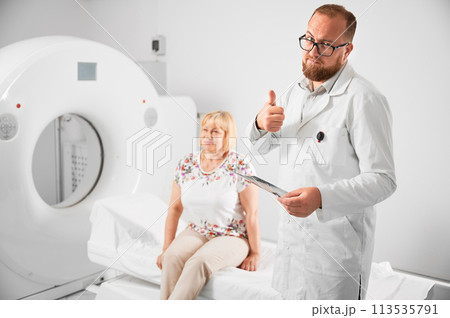 Medical computed tomography or MRI scanner. Doctor holding results of MRI, showing thumbs up gesture. Woman patient sitting on couch. Concept of medicine, healthcare and modern diagnostics. 113535791