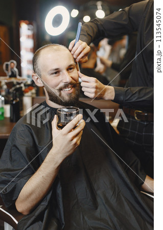 Man sitting in a chair. Hairdresser with a client. Guy drinkig a whiskey. Man sitting in a chair. Hairdresser with a client. Guy drinkig a whiskey. 113545074