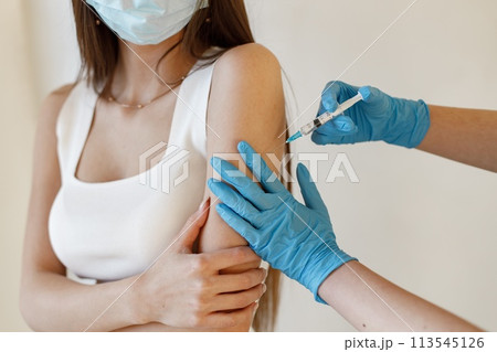 Young woman wearing mask and receiving coronavirus vaccine. A young girl receiving an immunity shot in the forearm. Closeup of doctor in blue gloves giving Covid-19, AIDS or flu antivirus vaccine to Young woman wearing mask and receiving coronavirus vaccine. A young girl receiving an immunity shot in the forearm. Closeup of doctor in blue gloves giving Covid-19, AIDS or flu antivirus vaccine to 113545126