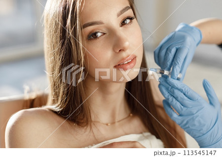 Closeup of doctor's hands in gloves with syringe near female face. Portrait of beautiful caucasian woman with white towel on her bodu. Girl receiving beauty injection. Closeup of doctor's hands in gloves with syringe near female face. Portrait of beautiful caucasian woman with white towel on her bodu. Girl receiving beauty injection. 113545147
