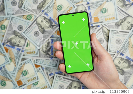 Hand holding smartphone with green screen on pile of dollar banknotes, top view 113550905