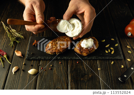 Close-up of a chef hands using a spoon adding sour cream to fried cutlets on a sorting board. The concept of preparing delicious food in a hotel 113552024