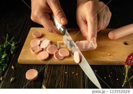 The cook cuts vienna sausage on a wooden cutting board. Preparing delicious sandwiches for breakfast on the kitchen table in a restaurant 113552029