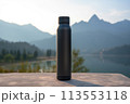 A black water bottle rests on a table against a backdrop of majestic mountains. The serene natural landscape features a clear blue sky and a peaceful lake in the distance 113553118