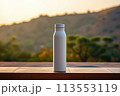 A plastic bottle filled with liquid is placed on a wooden table against the backdrop of a majestic mountain and clear blue sky, surrounded by lush green grass 113553119