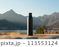 A black water bottle is placed on a rock overlooking a stunning natural landscape with mountains in the background, near a serene lake reflecting the blue sky 113553124
