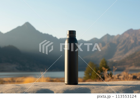 A black water bottle is placed on a rock overlooking a stunning natural landscape with mountains in the background, near a serene lake reflecting the blue sky A black water bottle is placed on a rock overlooking a stunning natural landscape with mountains in the background, near a serene lake reflecting the blue sky 113553124