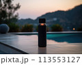 A black bottle rests on a wooden table overlooking a serene swimming pool, surrounded by lush plants with mountains in the horizon under a clear sky 113553127