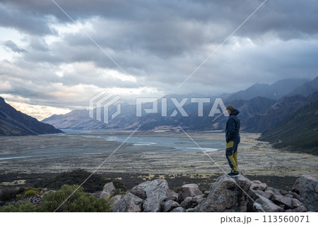 Male hiker looking down the alpine valley as sunset and storm are approaching it, New Zealand 113560071
