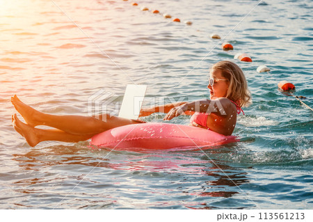 Woman works on laptop in sea. Freelancer, young blond woman in sunglases floating on an inflatable big pink donut with a laptop in the sea at sunset. Freelance, travel and holidays concept. 113561213