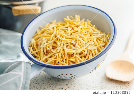 Uncooked spaetzle pasta in colander on kitchen table. Uncooked spaetzle pasta in colander on kitchen table. 113568853