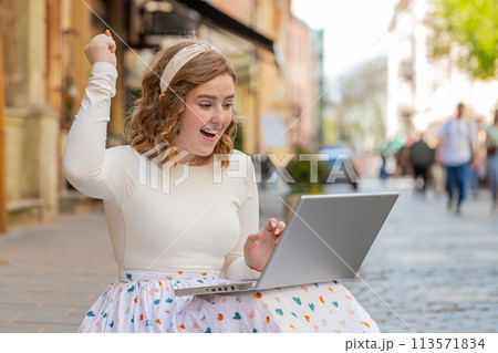 Caucasian happy woman working on laptop celebrate success win money sitting on urban street in city 113571834
