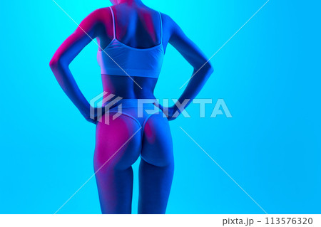 Rear view photo of toned female body, buttocks in neon pink light against gradient blue background. Model in lingerie put hands on hips. 113576320