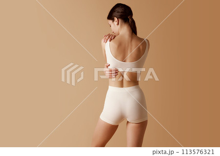 Rear view of slender woman in white sports top and shorts holding hands behind back against pastel beige background. Rear view of slender woman in white sports top and shorts holding hands behind back against pastel beige background. 113576321