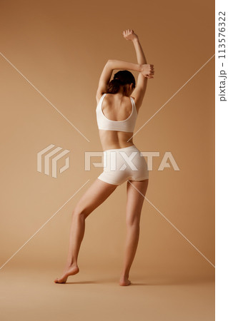 Rear view photo of young woman posing in sportswear that emphasizes her perfect curves against pastel beige background. Rear view photo of young woman posing in sportswear that emphasizes her perfect curves against pastel beige background. 113576322