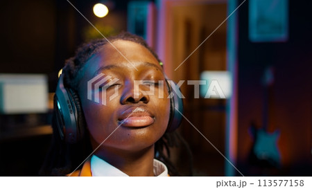 College student sitting laid back after working on class project, relaxing with ambience music on headphones at home desk. African american girl chilling with modern tunes on headset. Camera B. College student sitting laid back after working on class project, relaxing with ambience music on headphones at home desk. African american girl chilling with modern tunes on headset. Camera B. 113577158