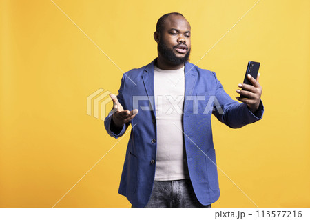 Annoyed man having unpleasant conversation with friends during teleconference meeting using smartphone, studio background. Hostile person having uncivil chat with mates during online videocall 113577216