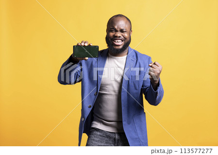 African american man excitedly celebrating while presenting isolated screen phone, studio background. Ecstatic BIPOC person holding copy space mockup cellphone used for advertising brands 113577277