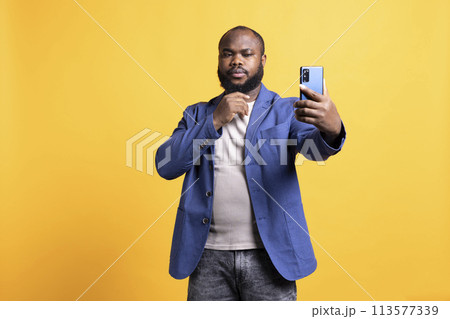 Narcissistic man using cellphone to take selfies, stroking his chin. Vain social media user taking photos using phone selfie camera, isolated over yellow studio background 113577339