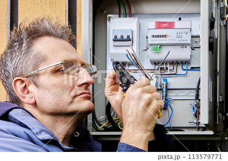 An electrician strips insulation from wire of three-phase power cable before connecting it to circuit breaker in an outdoor electrical panel. An electrician strips insulation from wire of three-phase power cable before connecting it to circuit breaker in an outdoor electrical panel. 113579771
