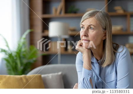 A serene mature woman in a blue shirt stares thoughtfully while sitting in a well-lit living room. The peaceful atmosphere highlights her pensive mood. A serene mature woman in a blue shirt stares thoughtfully while sitting in a well-lit living room. The peaceful atmosphere highlights her pensive mood. 113580331