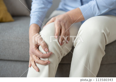 Senior woman sits in a living room, gently holding her sore knee, depicting common age-related health issues and the need for elderly care. Senior woman sits in a living room, gently holding her sore knee, depicting common age-related health issues and the need for elderly care. 113580343