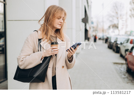 Beautiful smiling female with coffee cup using smartphone and standing outdoors. Phone Communication. Happy cheerful young woman walking on city street, Urban lifestyle concept. Traveler Beautiful smiling female with coffee cup using smartphone and standing outdoors. Phone Communication. Happy cheerful young woman walking on city street, Urban lifestyle concept. Traveler 113581217