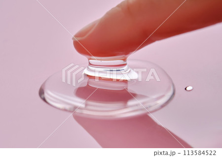 A finger touches a drop of cosmetic product on a pink background. 113584522