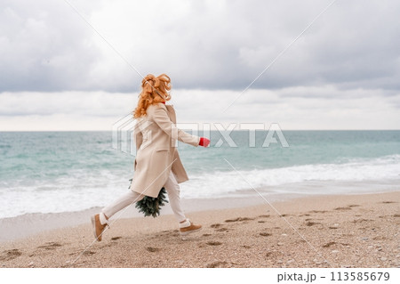 Redhead woman Christmas tree sea. Christmas portrait of a happy redhead woman walking along the beach and holding a Christmas tree in her hands. Dressed in a light coat, white suit and red mittens. 113585679