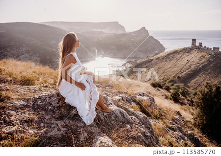 A woman in a white dress sits on a rock overlooking a body of water. The scene is serene and peaceful, with the woman enjoying the view and the calmness of the surroundings. 113585770