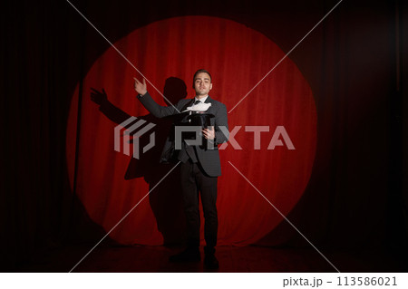 Magician with pigeon standing over red drapery in spotlight 113586021