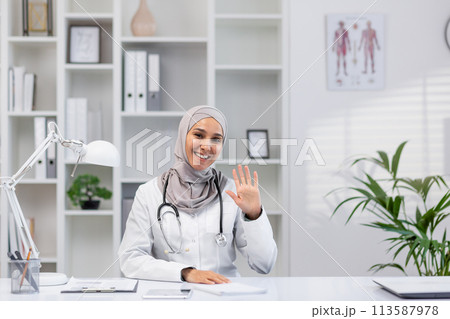 Smiling female doctor in hijab waves at the camera during a video call in a well-lit, modern office setting, displaying warmth and professionalism. Smiling female doctor in hijab waves at the camera during a video call in a well-lit, modern office setting, displaying warmth and professionalism. 113587978