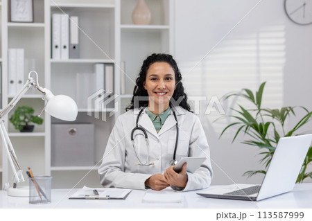 Confident female doctor with a stethoscope around her neck, smiling warmly in a well-lit, modern medical office, exuding professionalism and care. 113587999