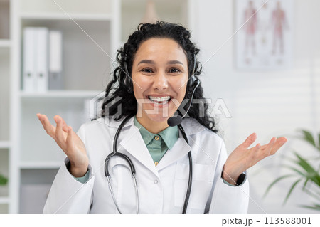 Cheerful female physician in a white coat using headphones for a video call. She looks into the camera with a friendly expression, offering medical advice and help. 113588001